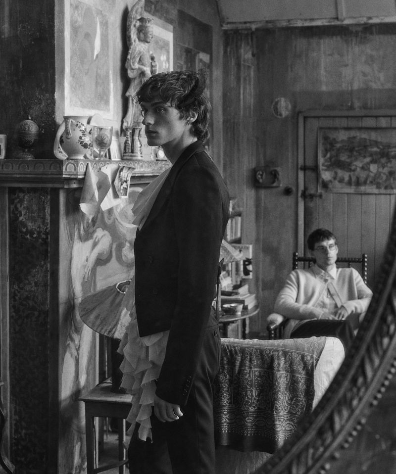 Black and white portrait of a man in a tailored jacket with ruffles standing in a room, with another man seated in the background.