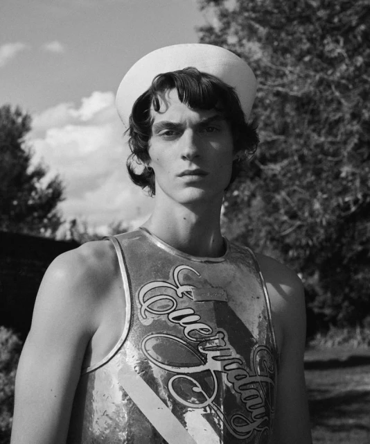 Black and white portrait of a young adult male model wearing a tank top and sailor hat in a natural, outdoor environment with trees.