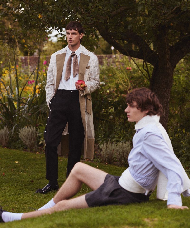 Two young adult men pose on a grassy lawn in stylish fashions, one standing and holding an apple, the other seated on the grass.