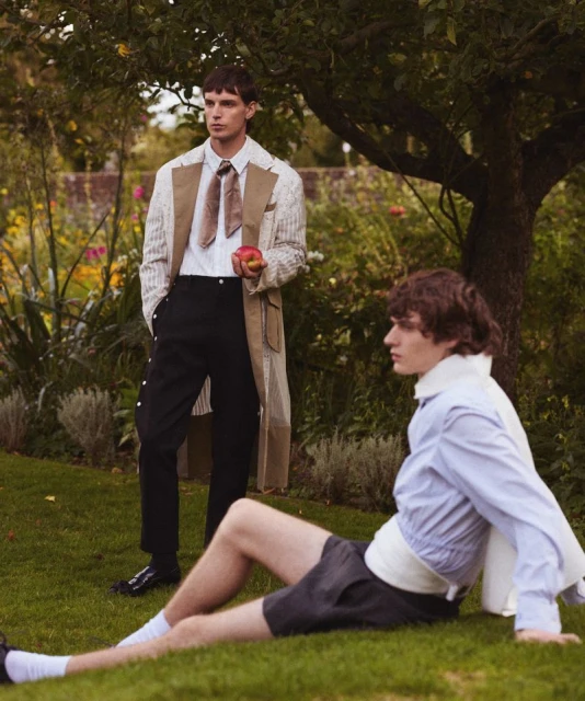 Two young adult men pose on a grassy lawn in stylish fashions, one standing and holding an apple, the other seated on the grass.