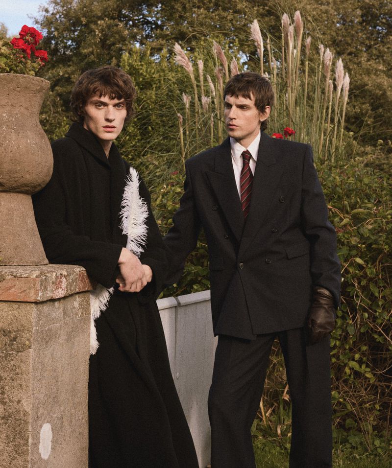Two male models posing in tailored dark coats and suits outdoors next to a stone urn and garden foliage.