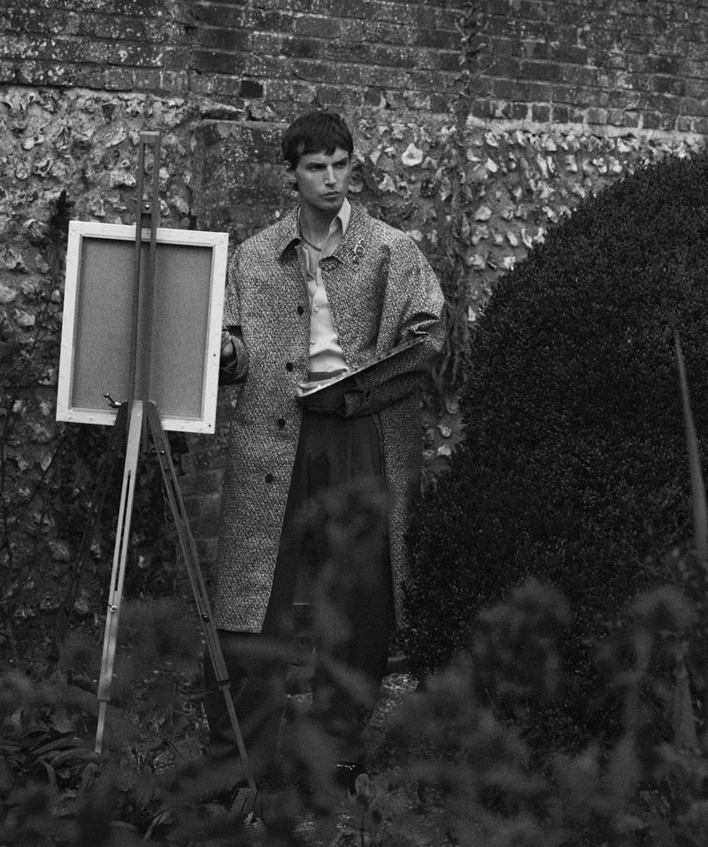 Young man with easel and palette in garden setting, wearing a coat. Black and white outdoor photography.