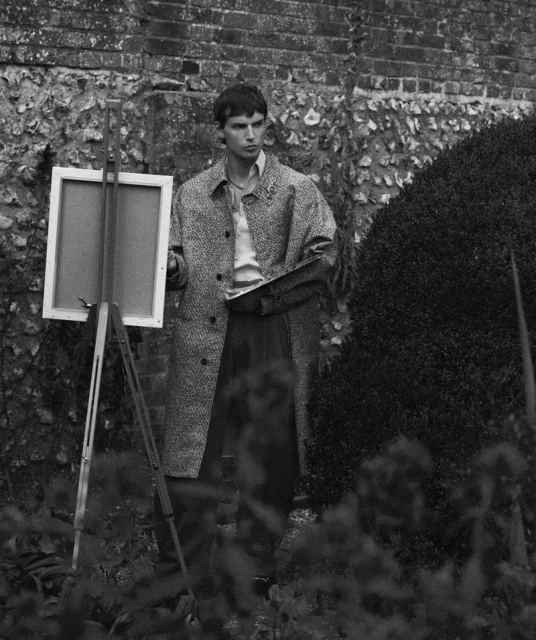 Young man with easel and palette in garden setting, wearing a coat. Black and white outdoor photography.