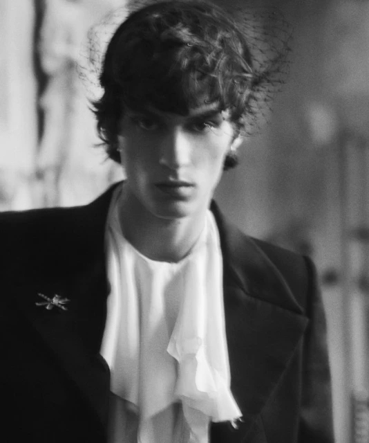 Monochromatic portrait of a person in a jacket and ruffled shirt, accessorized with a vintage brooch and decorative netting over hair.