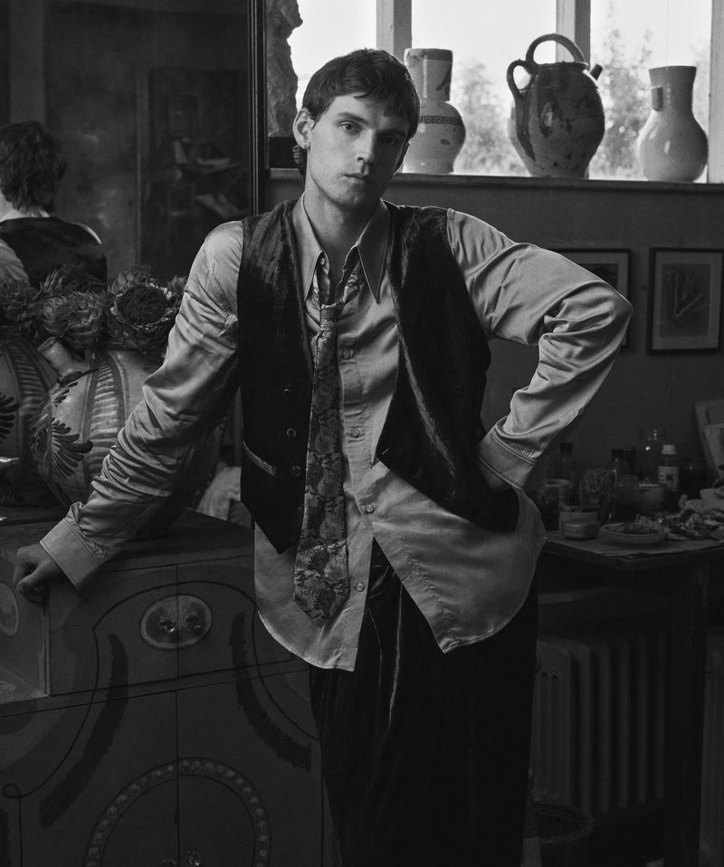 Monochrome portrait of a young man indoors wearing a vest and patterned tie, posing with hand on an ornate cabinet.