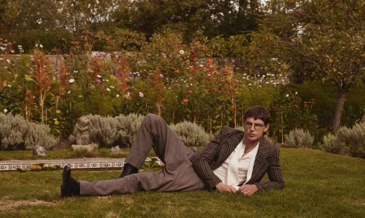Model in a striped blazer and grey trousers lying on a lawn in front of a lush garden for HERO Magazine