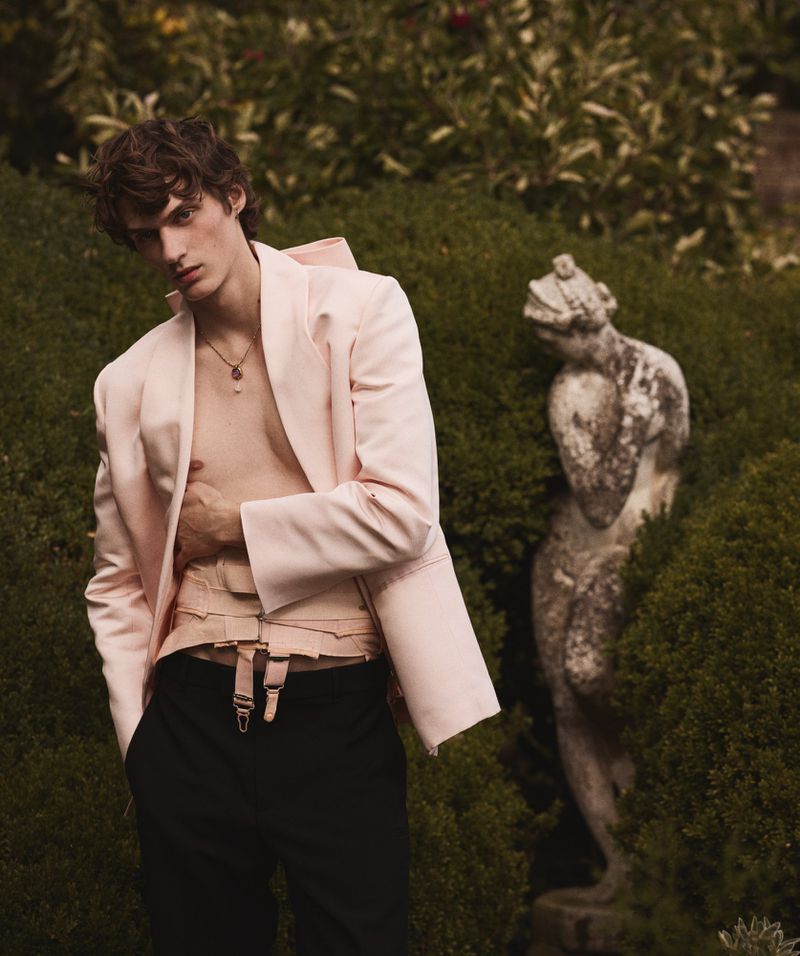 Stylish man poses in a pink blazer and black pants in an outdoor garden setting with lush green foliage and a stone statue.