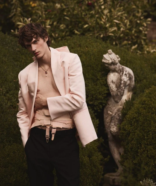 Stylish man poses in a pink blazer and black pants in an outdoor garden setting with lush green foliage and a stone statue.