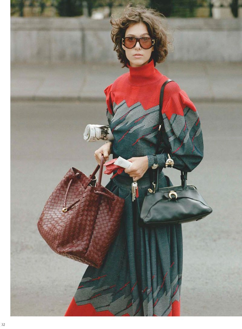 Model Aya Fellaini wearing a red and teal patterned knit dress and oversized sunglasses while holding two bags on a city street.