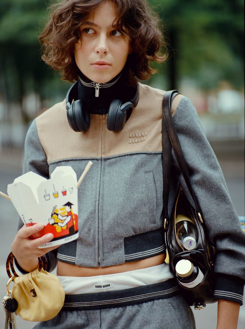 Woman in gray tracksuit and jacket with headphones around neck, holding takeout container in an urban outdoor setting.