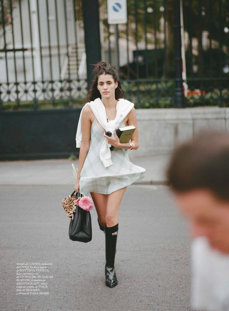 A young woman in a light dress and black boots walks along a city street carrying a black bag and a book in an eye-level shot.