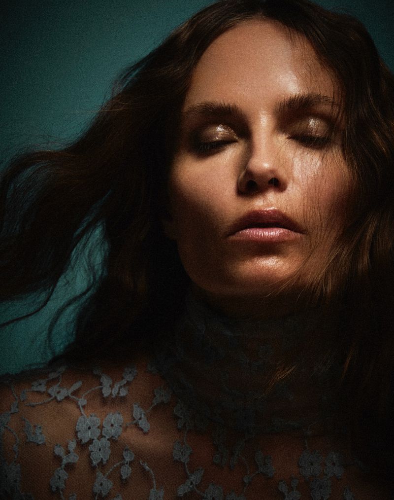 Portrait of a woman with long brown hair and closed eyes, wearing a blue lace top against a teal background in soft, diffused lighting.