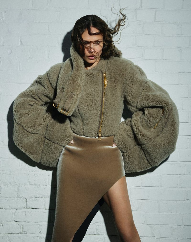 Woman poses in a studio wearing a furry jacket and a skirt with a slit. She stands against a white brick wall with soft lighting.