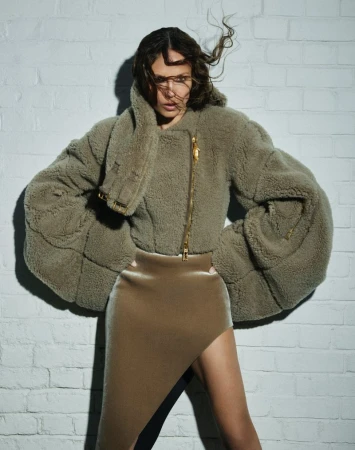Woman poses in a studio wearing a furry jacket and a skirt with a slit. She stands against a white brick wall with soft lighting.