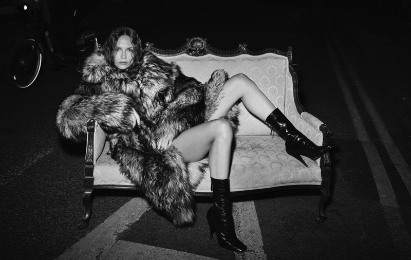 A woman in a fur coat and boots poses on a sofa on a street at night. The black and white portrait features a dark, moody atmosphere.