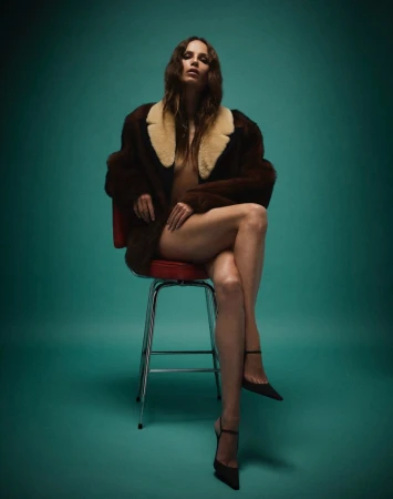 Woman in a brown faux fur coat poses while seated on a red stool in a studio with a teal backdrop and wearing strappy heels.