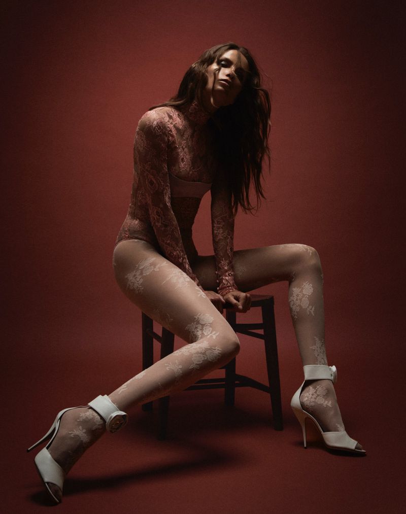 A slender brunette woman poses gracefully in a lace bodysuit, tights, and high heels against a deep red backdrop, seated on a simple stool.