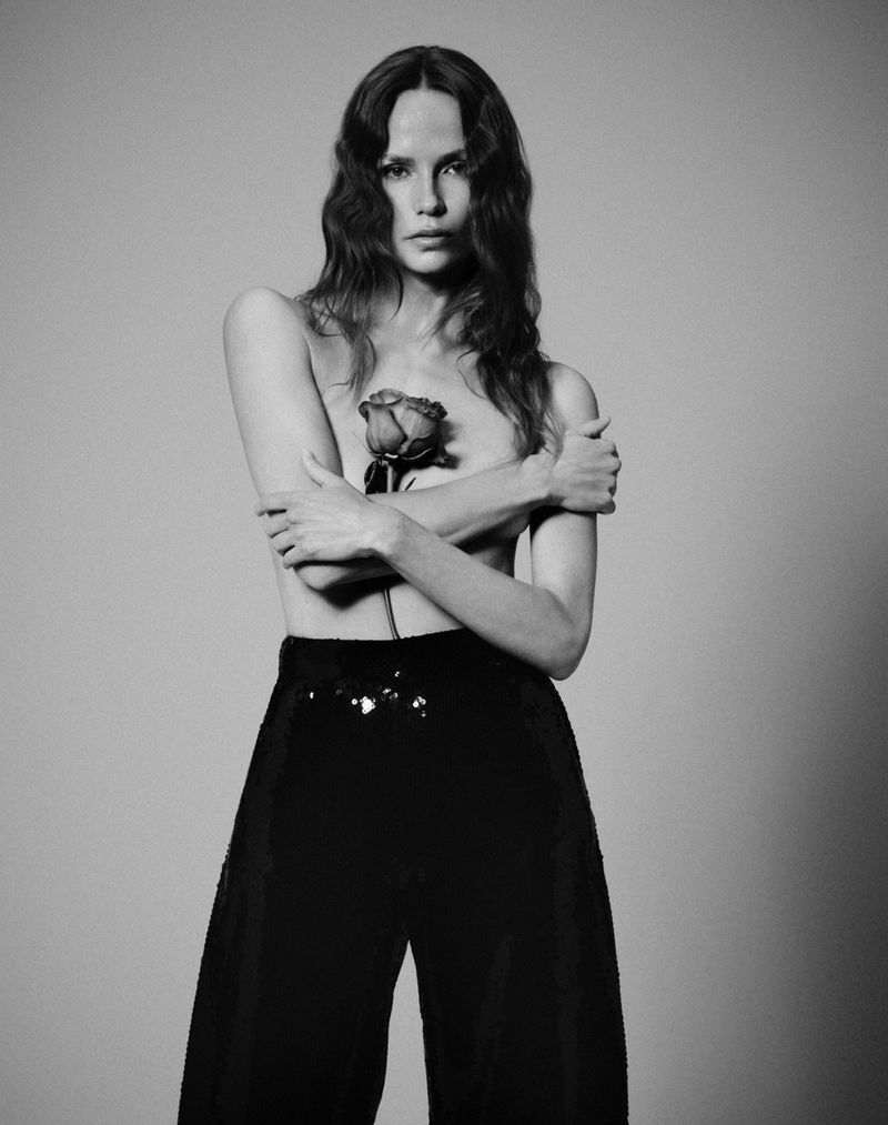 Monochrome portrait of a slim adult woman with long curly hair, bare shoulders, and black pants holding a rose in a neutral studio.