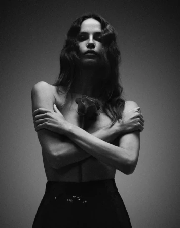A portrait in grayscale of a young adult woman with long wavy hair holding a single red rose in a photo studio setting.