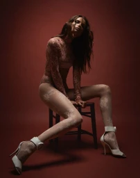 Model Natasha Poly seated on a stool wearing a sheer floral lace bodysuit, matching lace tights, and white stiletto heels on a red backdrop.
