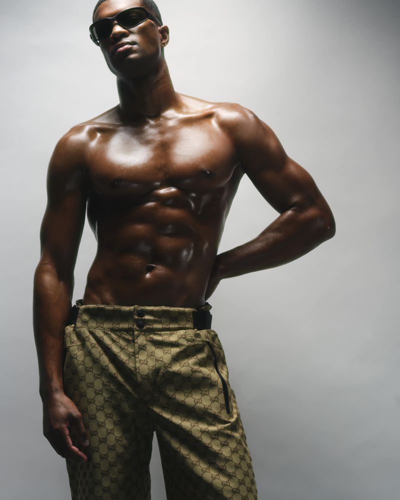 Shirtless African American male model in Gucci pants and sunglasses strikes a pose in a bright photo studio, fashion-forward.