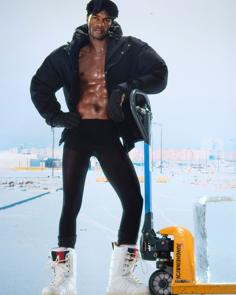 Shirtless man in a puffy jacket, goggles, and ski boots poses next to a forklift with an ice block, set in an industrial outdoor area.