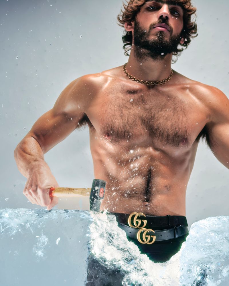 Athletic man shirtless breaking ice block with a hammer, wearing a gold chain and a Gucci belt against a light background.