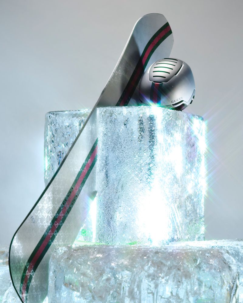 Snowboard and helmet with Gucci logo on ice block against a light background in a studio. The snowboard has a stripe design.