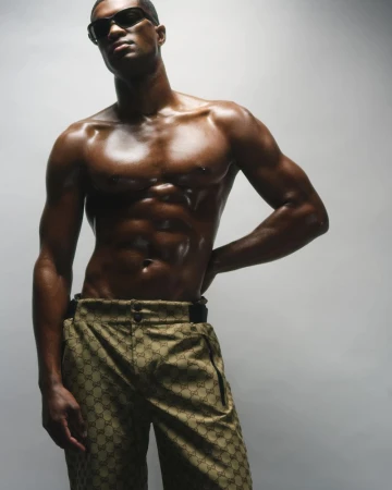 Shirtless male model wearing dark sunglasses and patterned trousers against a grey backdrop
