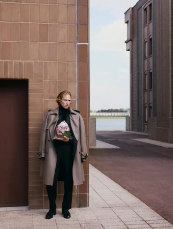 A fair-skinned person in a tan trench coat stands on a city street holding a clear plastic terrarium with pink flowers and green plants.