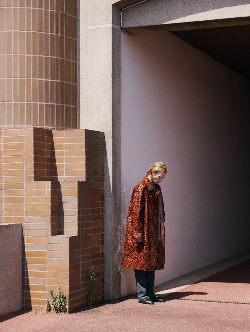 Adult fashion model in a patterned orange coat, grey pants, and black shoes stands near a concrete wall with a geometric brick design.