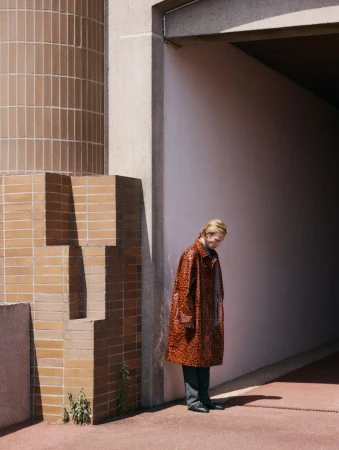 Adult fashion model in a patterned orange coat, grey pants, and black shoes stands near a concrete wall with a geometric brick design.