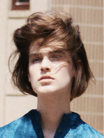 Close-up studio portrait of a young adult female with wavy auburn hair, wearing a blue patterned blouse and posing against a neutral background.