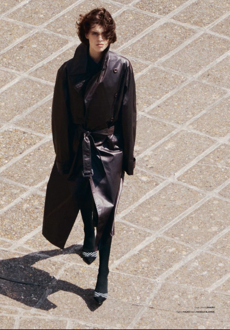 A woman in a dark leather trench coat walks on a tan stone-paved surface, featuring long sleeves, a belted waist, and high-heeled pumps.