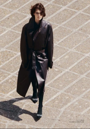 A woman in a dark leather trench coat walks on a tan stone-paved surface, featuring long sleeves, a belted waist, and high-heeled pumps.