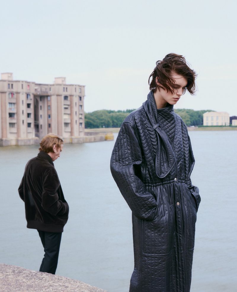 A young adult woman in a black quilted coat poses outdoors near a river, with another person and a building in the background.