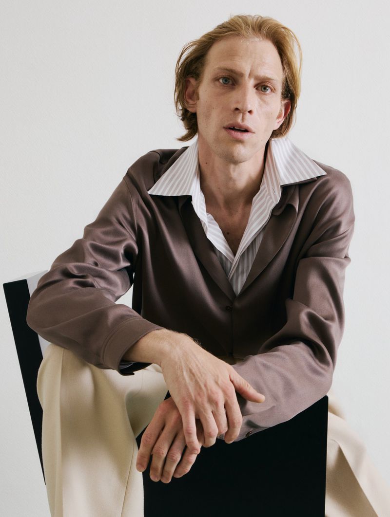Young adult man with fair skin and light brown hair poses in a studio wearing a brown jacket and beige pants while seated on a chair.