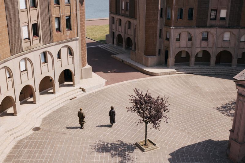 High-angle view of two adults walking across a plaza towards a leafless tree, surrounded by tall, arched buildings and a distant river.