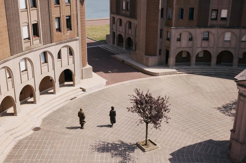 High-angle view of two adults walking across a plaza towards a leafless tree, surrounded by tall, arched buildings and a distant river.
