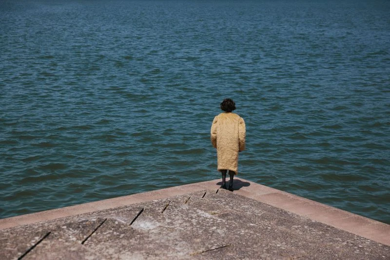 A woman in a yellow coat stands at the edge of a concrete structure overlooking a large body of water in a daytime scene.