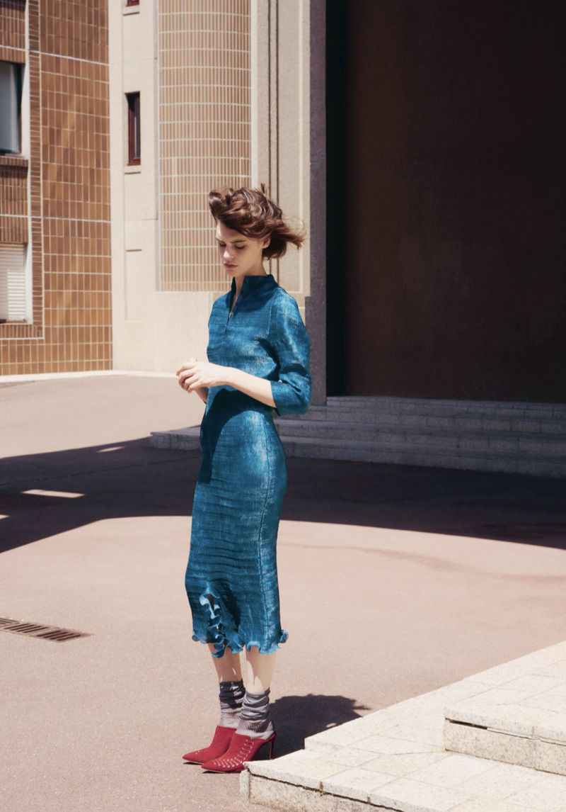 A stylish woman in a blue dress stands in front of a tan building, wearing patterned socks and red heeled boots on a sunny day.
