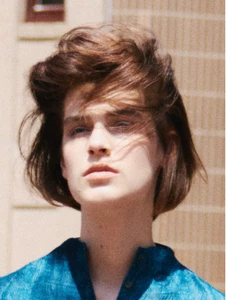 Close-up portrait of a model with voluminous wind-swept brown hair wearing a patterned blue shirt outdoors.