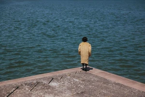 Model viewed from behind in a textured tan coat standing at the corner of a concrete pier overlooking rippling open water.