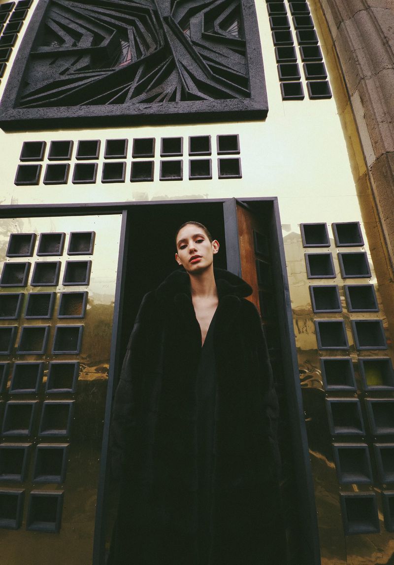 Model Valentina Navarrete posing in a long black coat within a doorway flanked by golden geometric architectural panels.