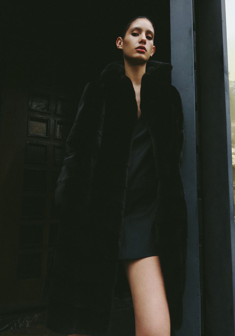 Fashionable woman in a black coat poses against a dark doorway in a dimly lit, urban environment.