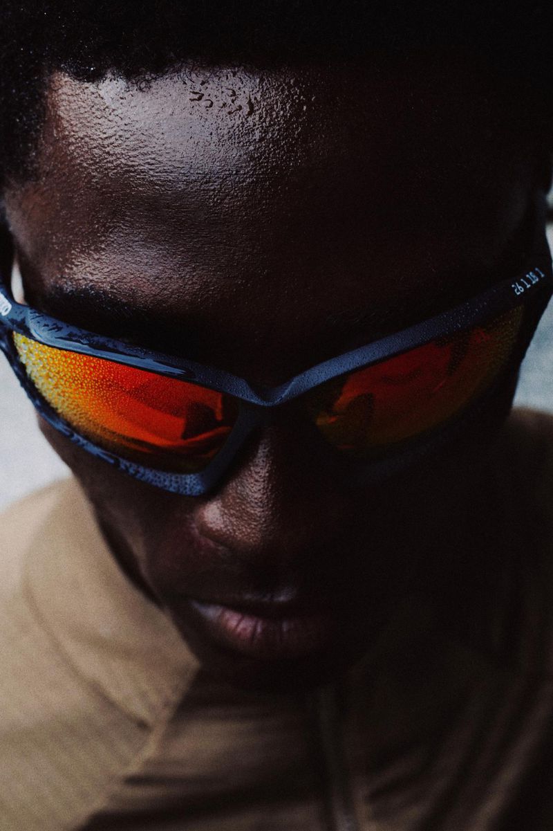 Close-up of a dark-skinned male wearing sporty goggles with orange lenses, illuminated by contrasting light, creating a portrait.