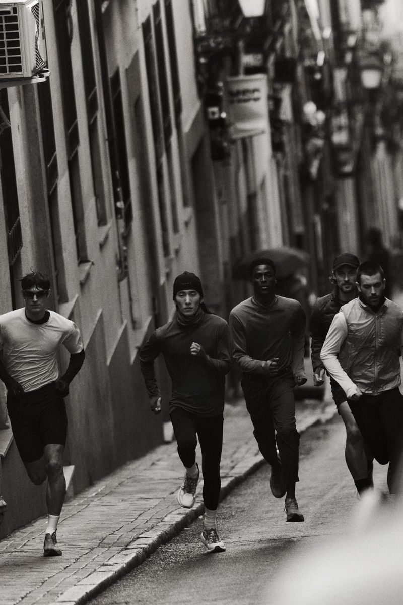 Black and white image captures a group of runners, including men of Asian and African descent, sprinting down a narrow city street.