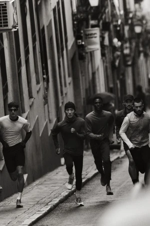 Black and white image captures a group of runners, including men of Asian and African descent, sprinting down a narrow city street.
