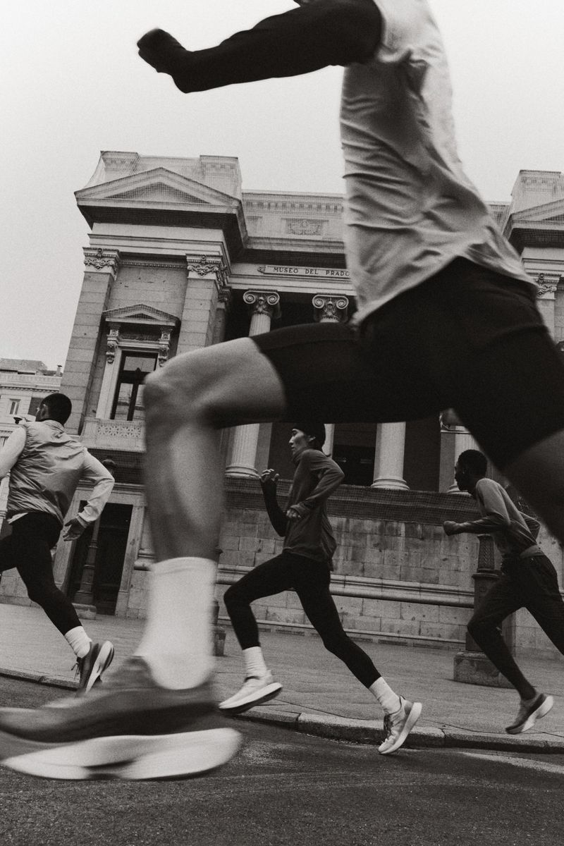 Four runners sprint along a city street past classical architecture in a black and white long exposure photograph.