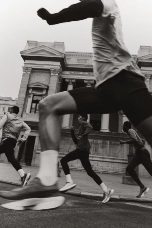 Four runners sprint along a city street past classical architecture in a black and white long exposure photograph.